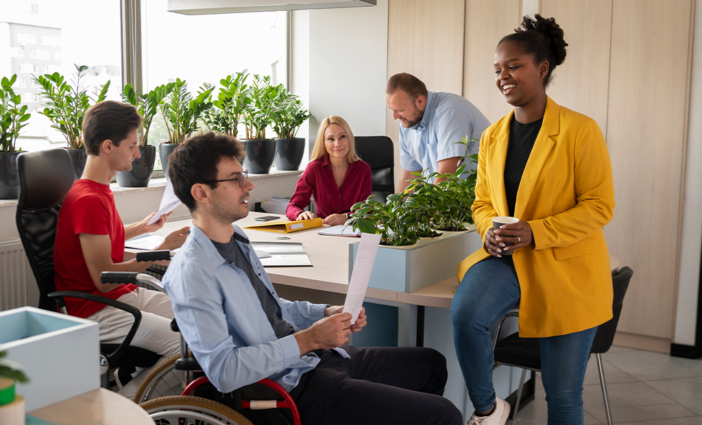 ROI da acessibilidade. Grupo diverso em reunião informal de trabalho em escritório moderno com plantas e luz natural.