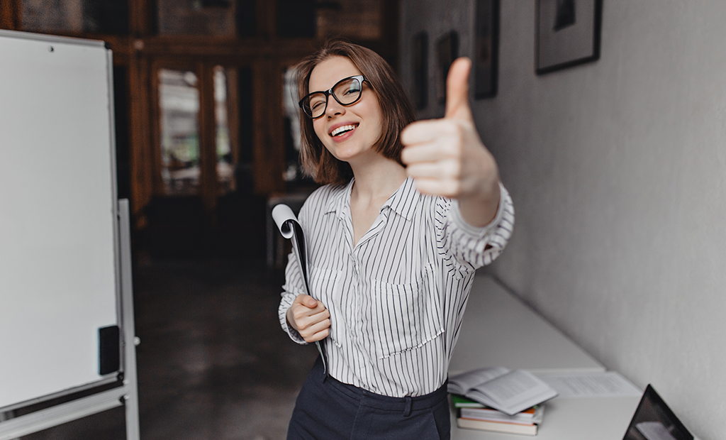ROI da acessibilidade. Mulher sorridente com óculos faz sinal de positivo em ambiente de trabalho com documentos ao fundo.
