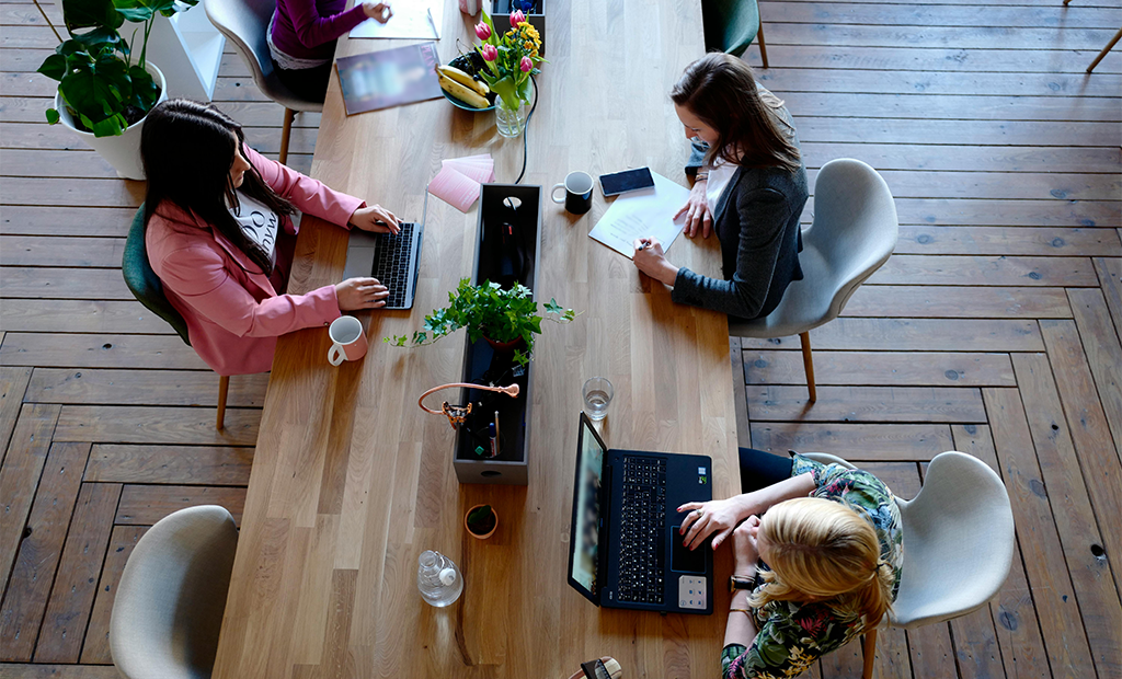 Acessibilidade digital. Três mulheres trabalhando em mesa de madeira com laptops e anotações, vistas de cima, em ambiente de escritório.
