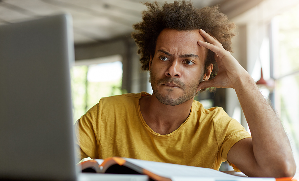 Acessibilidade digital. Homem de camiseta amarela e cabelo cacheado volumoso olha para tela de laptop com expressão preocupada.