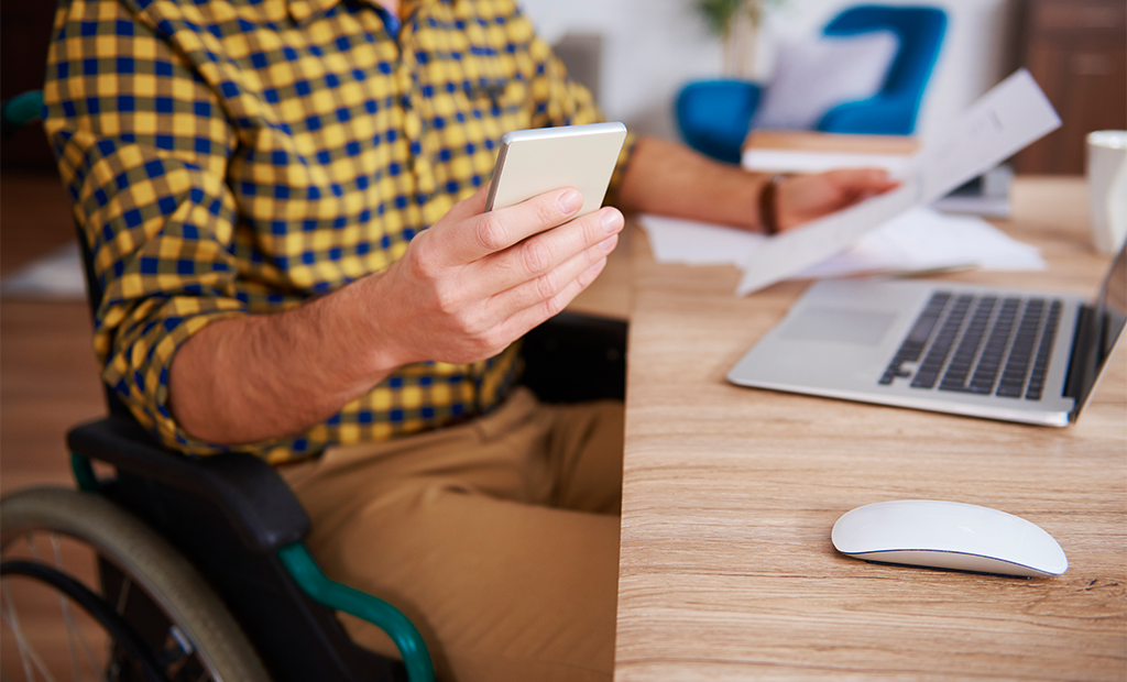 Acessibilidade digital. Homem em cadeira de rodas segura celular e documento, trabalhando em mesa com laptop em ambiente de escritório.
