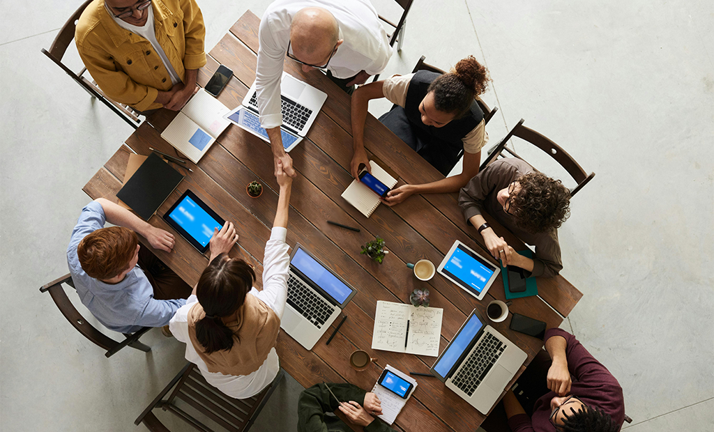 Acessibilidade digital. Grupo diverso reunido em mesa de madeira, com laptops e tablets, enquanto duas pessoas se cumprimentam com aperto de mão.