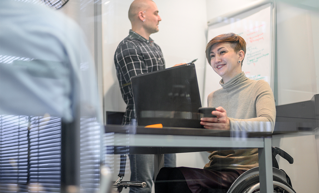 ESG. Mulher em cadeira de rodas sorri enquanto trabalha no notebook, com colega em pé escrevendo em quadro branco.