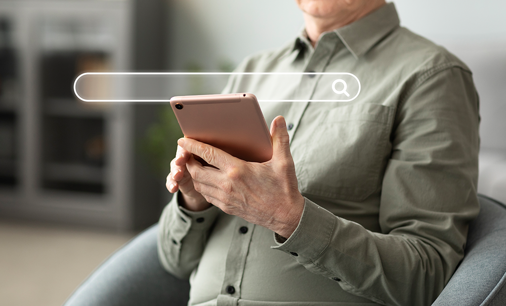 Pessoa idosa sentada em uma poltrona realiza uma pesquisa na internet utilizando um tablet.